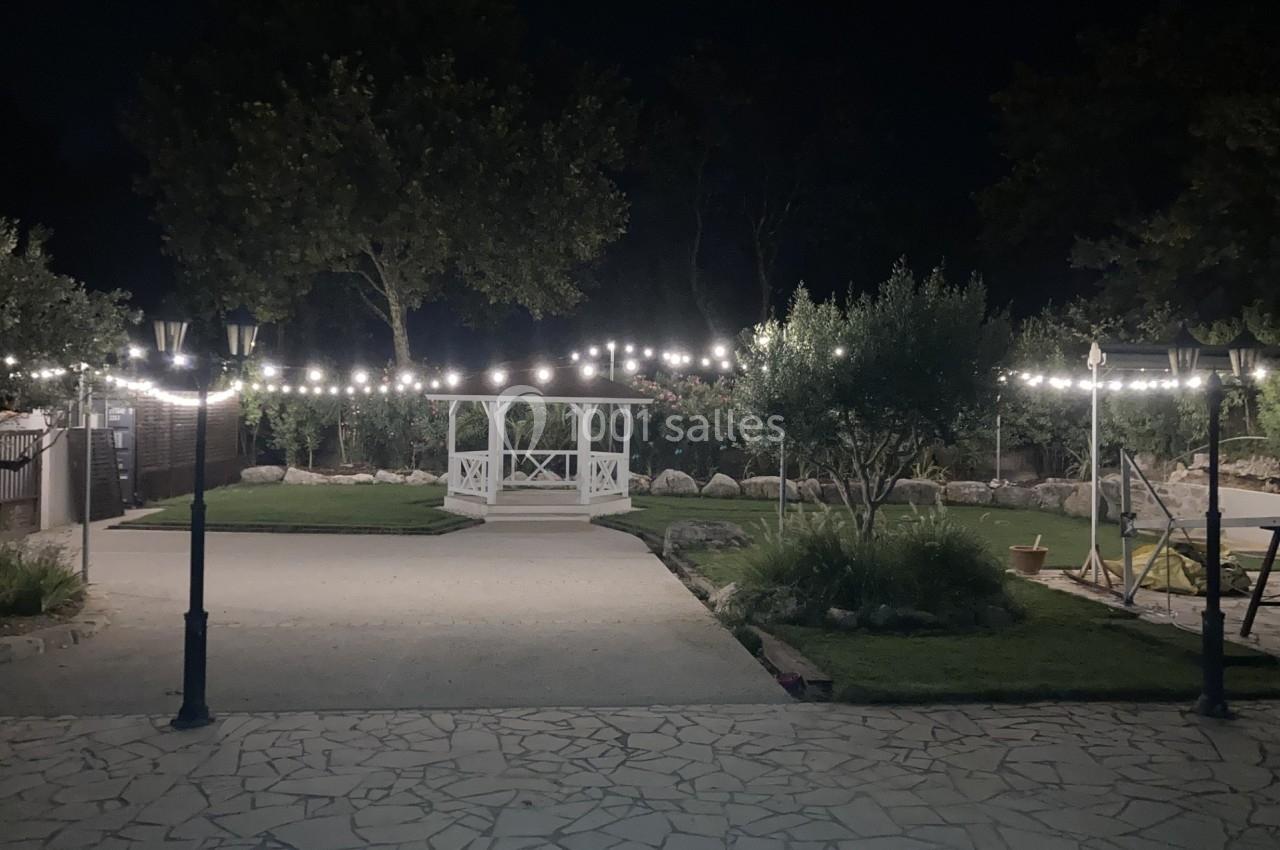 Allée pavée éclairée par des guirlandes lumineuses, menant à une pergola blanche dans un jardin nocturne.