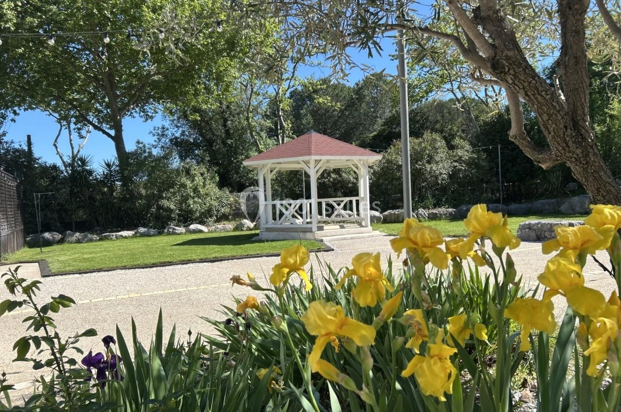 Kiosque blanc avec toit rouge dans un parc verdoyant, entouré de fleurs jaunes et d'arbres sous un ciel bleu.