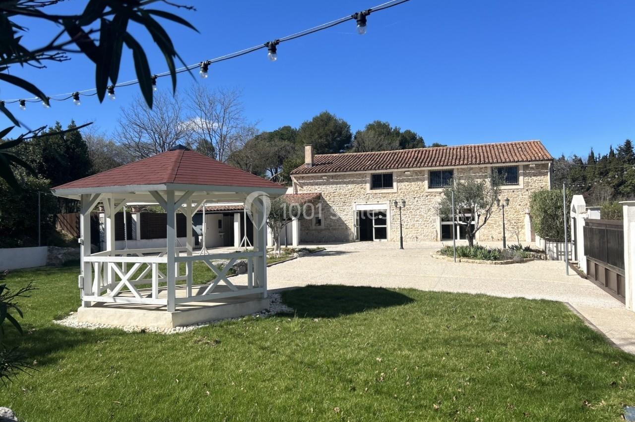 Maison en pierre avec toit en tuiles, jardin arboré, gazébo blanc et guirlandes lumineuses sous un ciel bleu.
