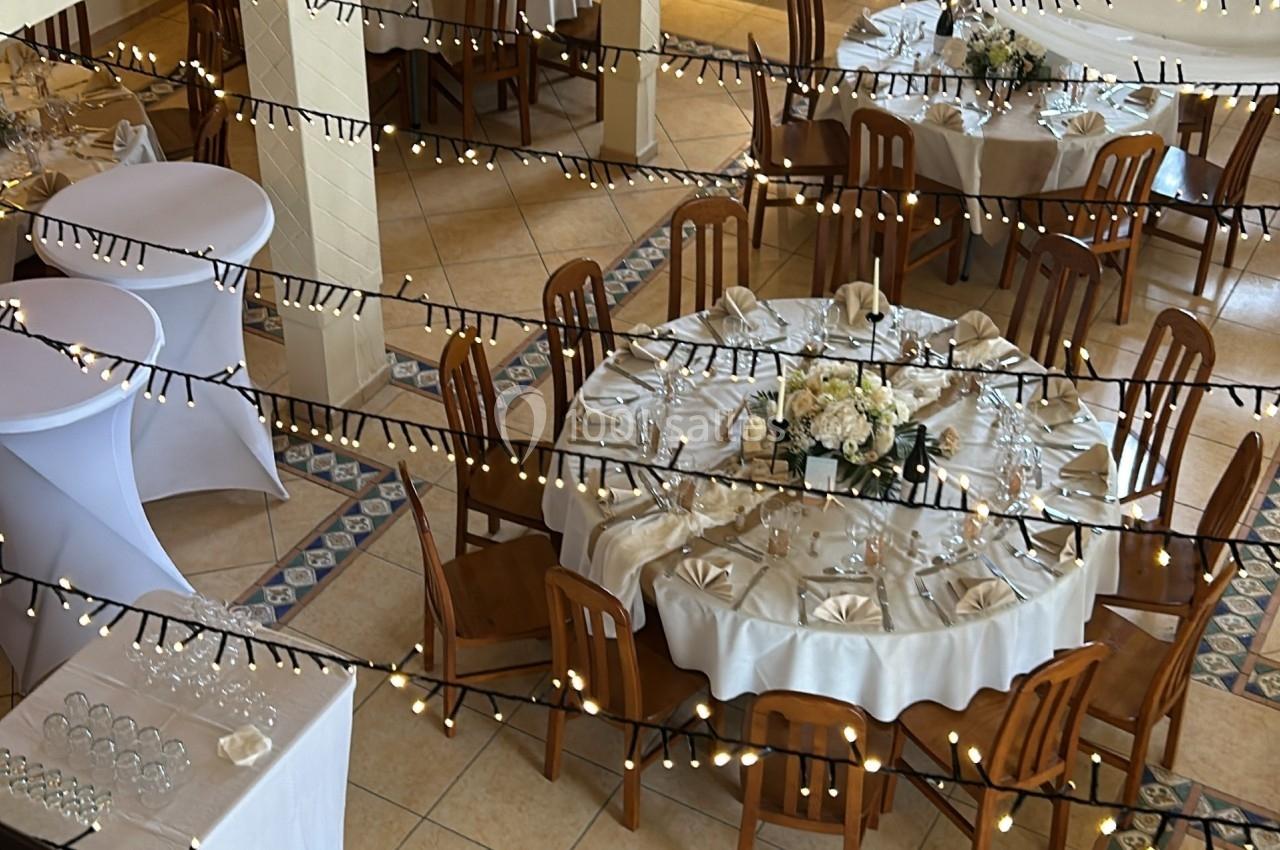 Salle décorée pour un événement avec tables rondes dressées, nappes blanches, chaises en bois et guirlandes lumineuses…