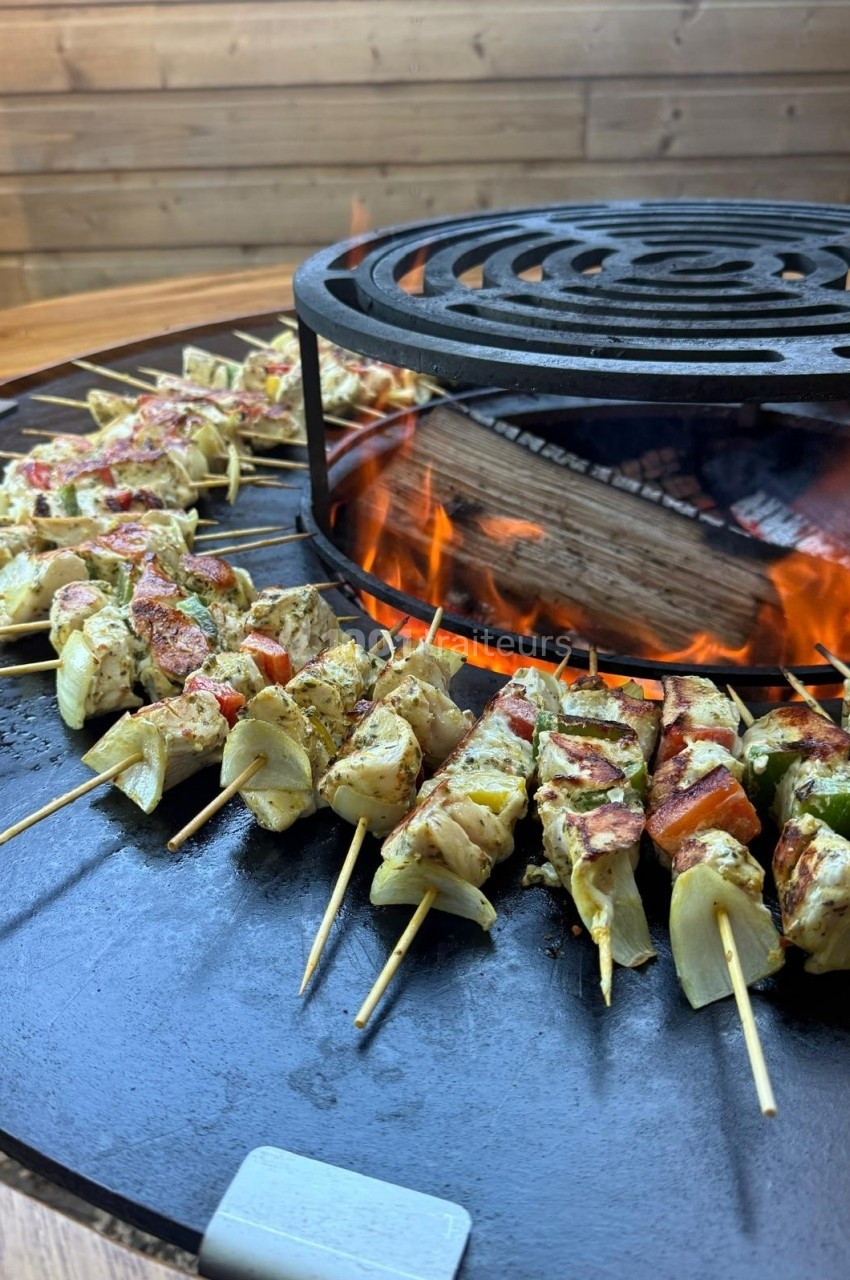 Brochettes de légumes et de viande grillant sur une plancha circulaire avec un feu de bois au centre.