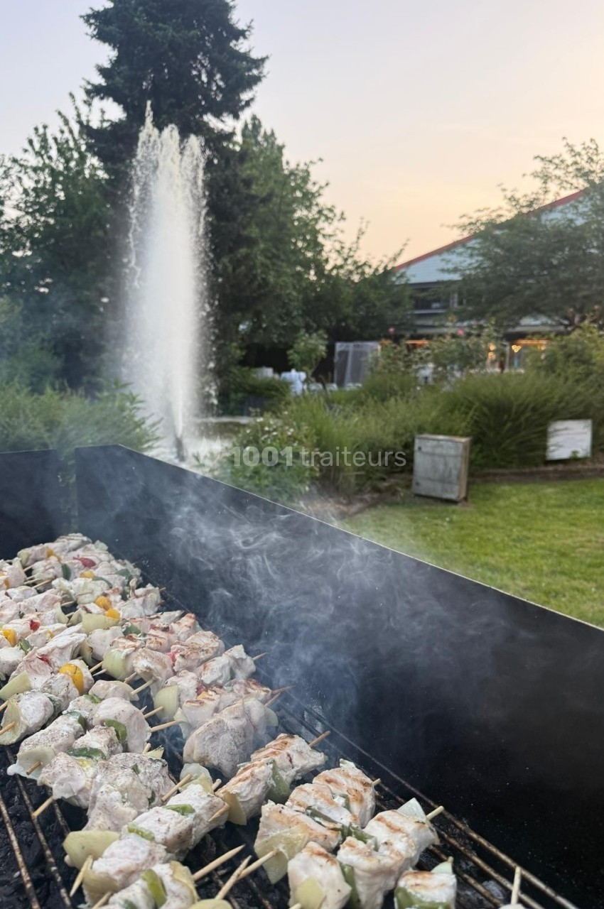 Brochettes de viande et légumes grillant sur un barbecue en plein air, avec une fontaine et un jardin en arrière-plan.