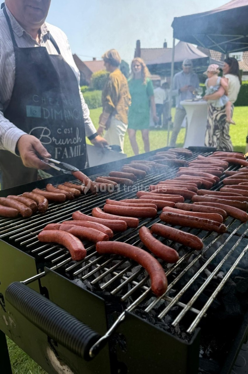 Un homme fait griller des saucisses sur un barbecue lors d'un événement en plein air avec des invités en arrière-plan.
