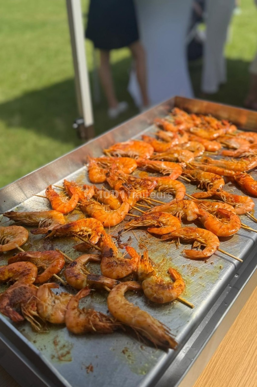Crevettes grillées sur une plaque de cuisson en extérieur, avec des personnes floues en arrière-plan sur une pelouse.