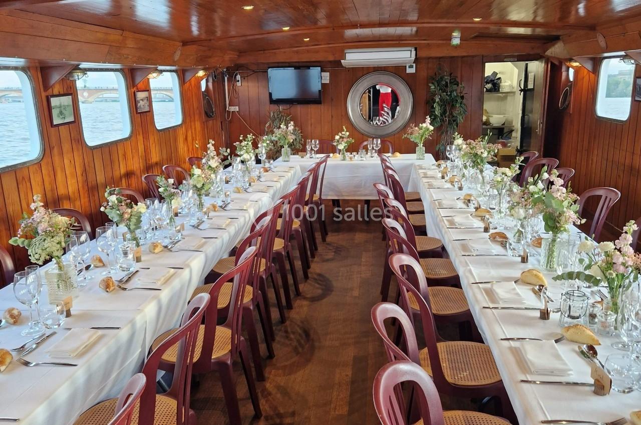 Salle de réception sur un bateau avec tables dressées, nappes blanches, chaises en bois et décorations florales.