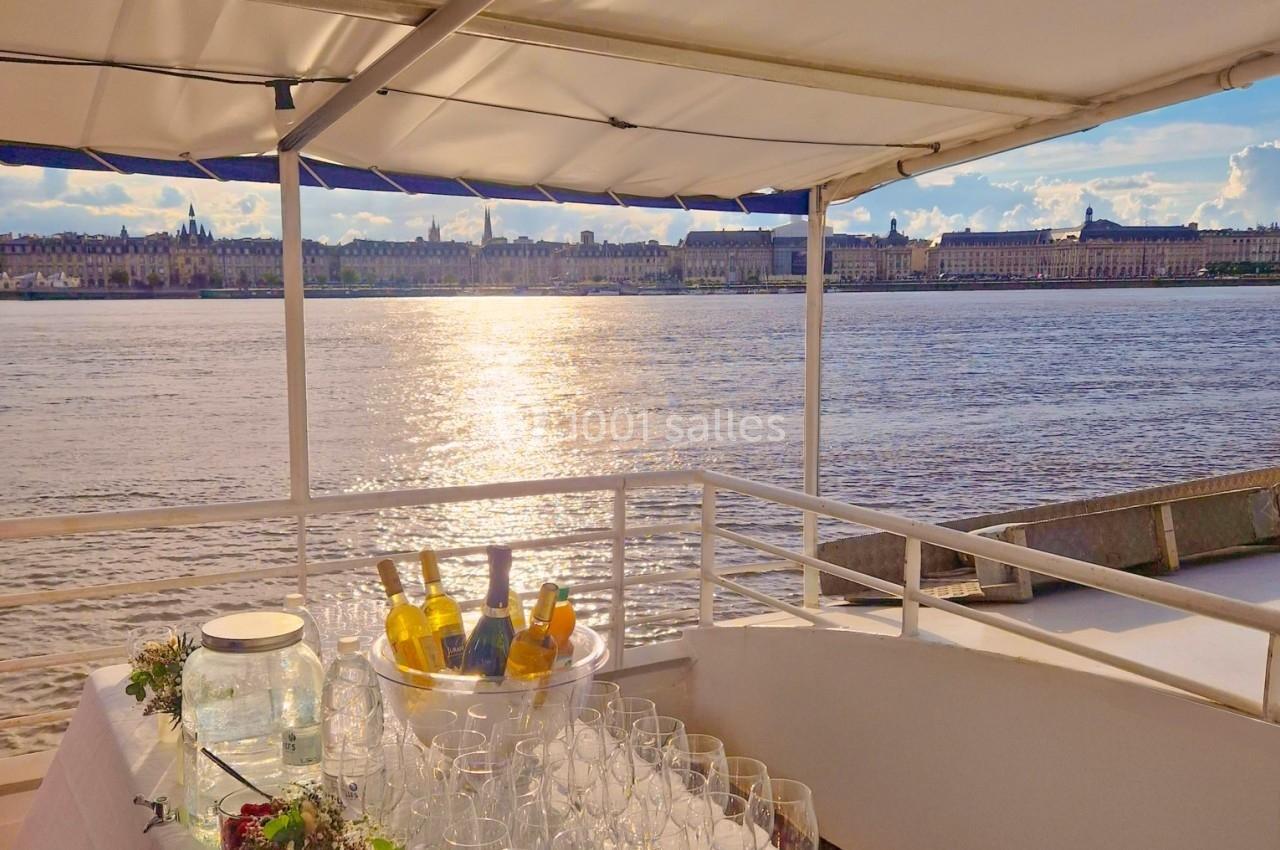 Table dressée avec des verres, carafes et fleurs, sur un bateau avec vue sur un fleuve et une ville en arrière-plan.