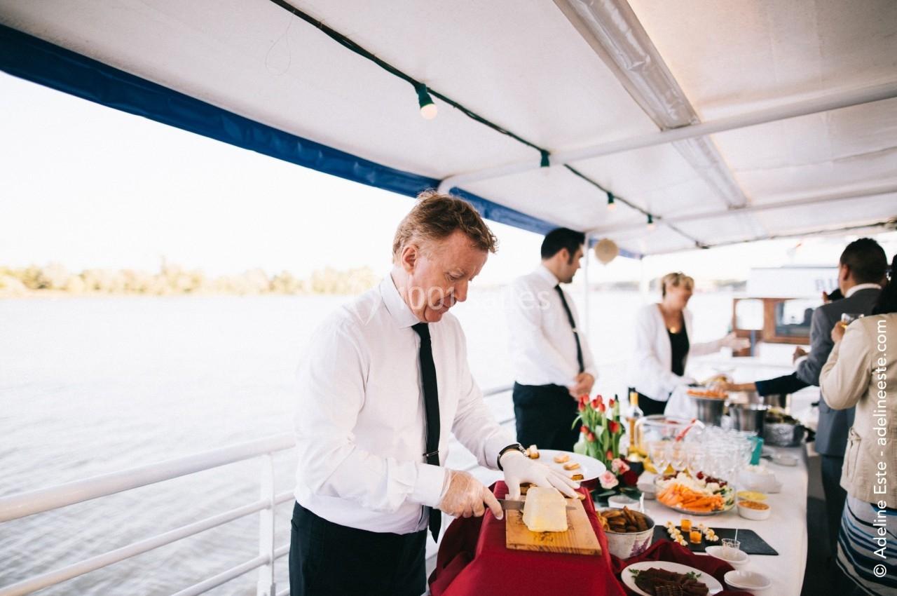 Un serveur en chemise blanche découpe du fromage sur une table garnie de mets, à bord d'un bateau.
