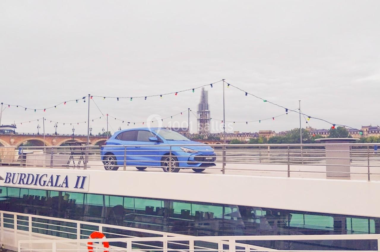 Une voiture bleue est exposée sur le pont supérieur d'un bateau amarré, avec une ville et un clocher en arrière-plan.