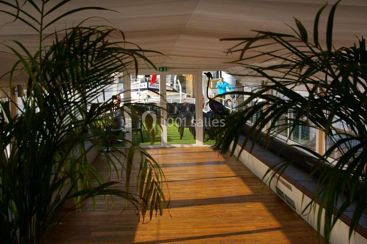 Vue d'un couloir lumineux avec un sol en bois, bordé de plantes vertes et menant à une porte vitrée donnant sur l'extérieur.