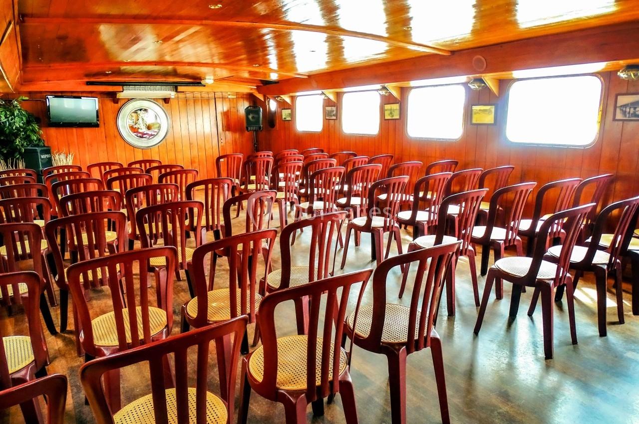 Salle en bois avec des rangées de chaises vides en plastique et assises en osier, éclairée par des fenêtres latérales.