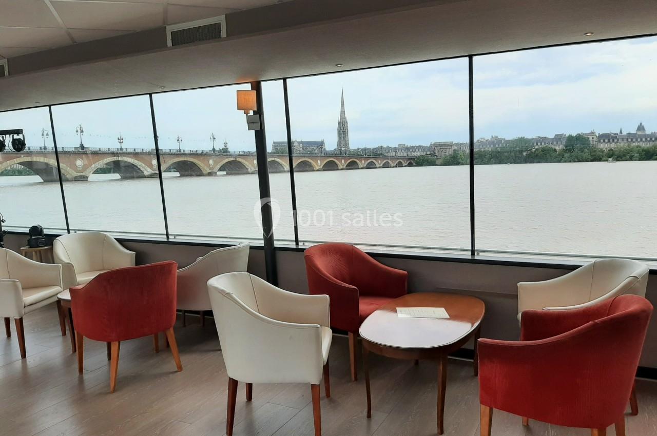 Salon avec fauteuils rouges et blancs, tables en bois, et vue sur un fleuve, un pont et une église à l'extérieur.