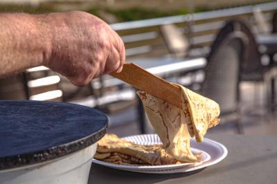 Une main dépose une crêpe chaude sur une assiette à l'aide d'une spatule, près d'une crêpière en extérieur.