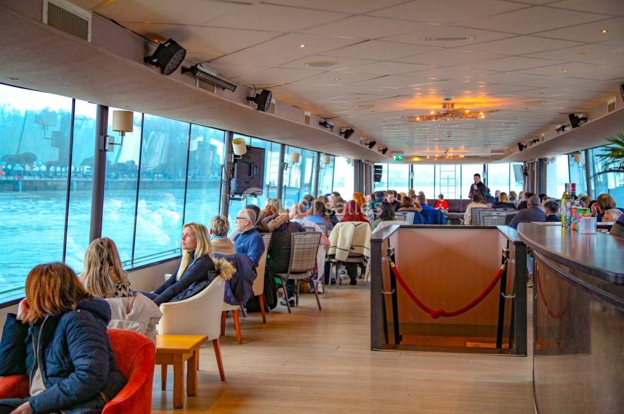 Intérieur d'un bateau avec des passagers assis près de grandes fenêtres donnant sur une rivière.