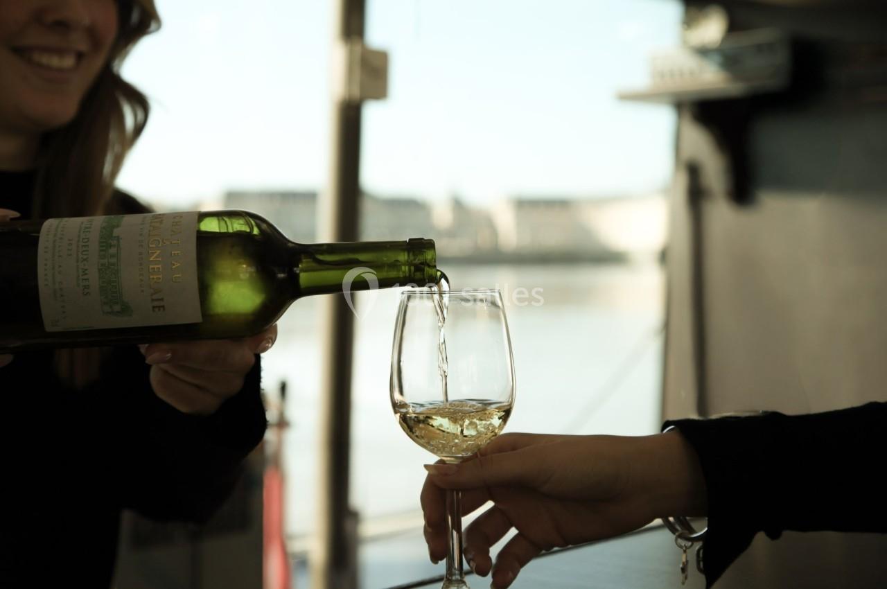 Une personne verse du vin blanc dans un verre tenu par une autre, avec une vue fluviale en arrière-plan.