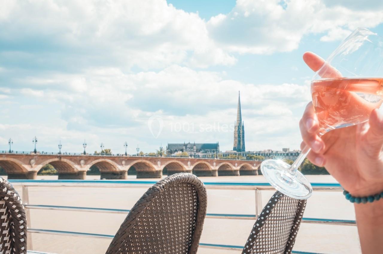 Une personne tient un verre de vin rosé en terrasse avec vue sur un pont et une église sous un ciel partiellement nuageux.