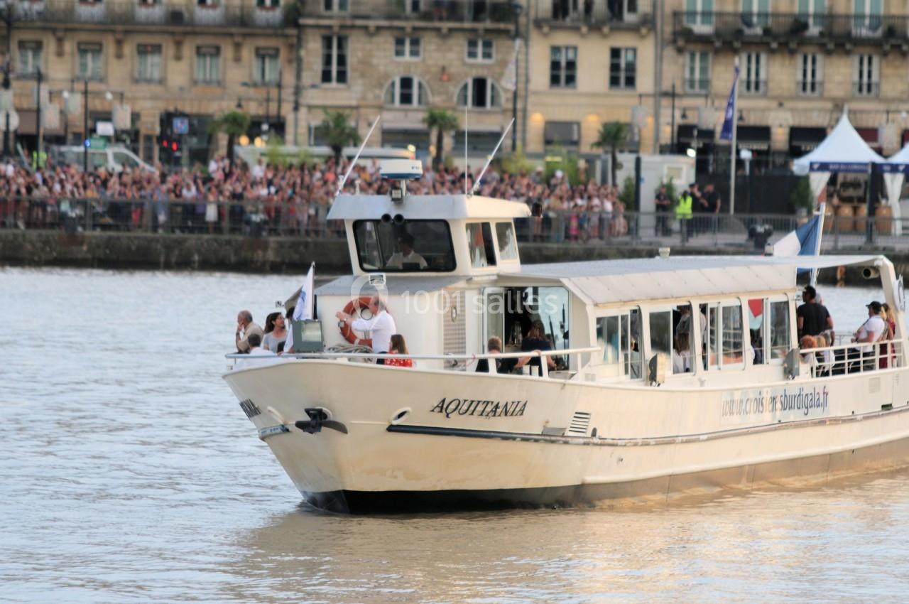 Bateau de tourisme nommé ’Aquitania’ naviguant sur une rivière, avec des passagers à bord et des spectateurs sur la rive.