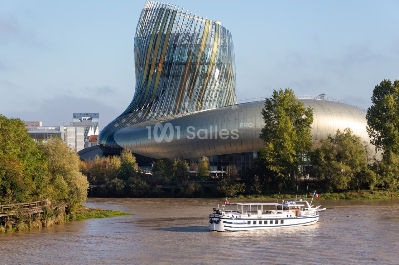 Bâtiment moderne en verre et métal au bord d'une rivière, avec un bateau blanc naviguant au premier plan.