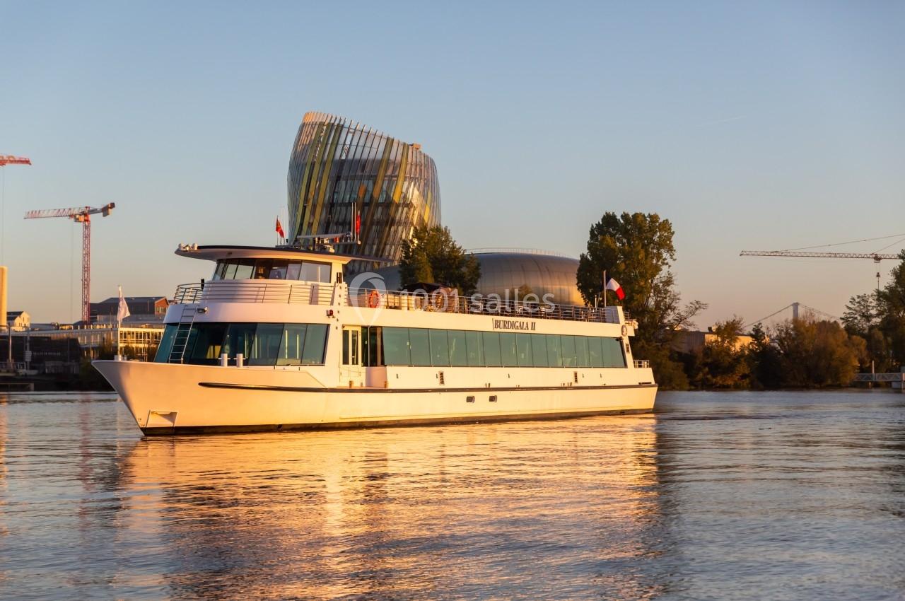 Bateau de croisière naviguant sur une rivière au coucher du soleil, avec un bâtiment moderne en arrière-plan.