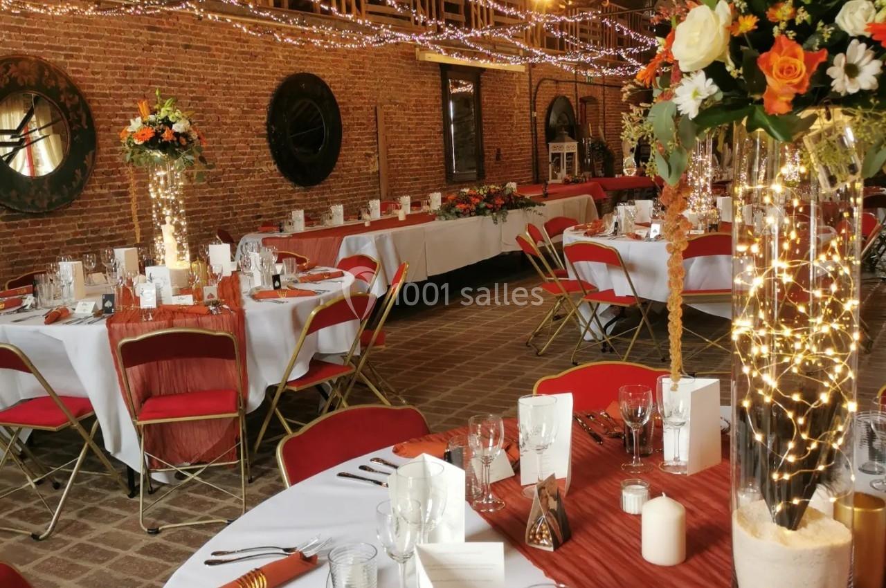 Salle de réception décorée avec des tables rondes, nappes blanches, chaises rouges et arrangements floraux lumineux.