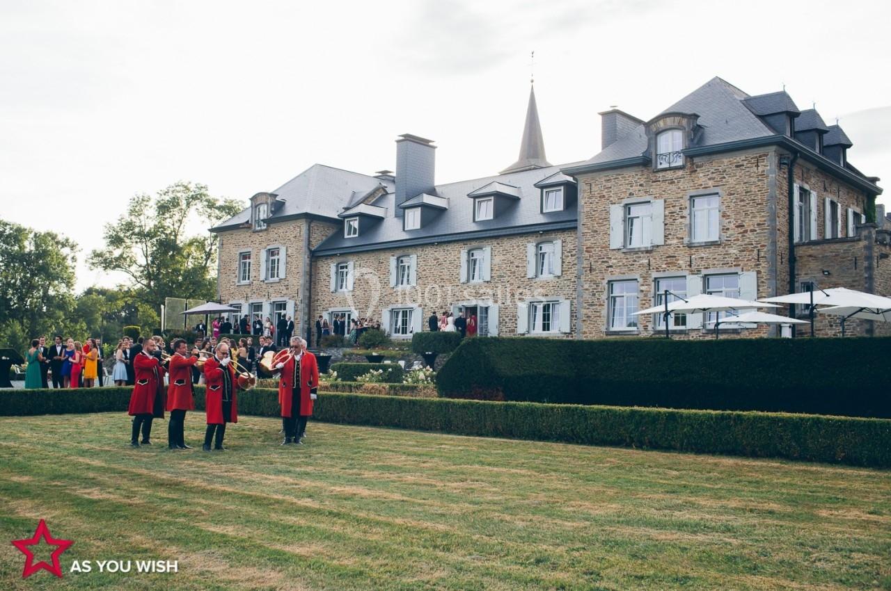 Un groupe de musiciens en tenue rouge joue devant un grand bâtiment en pierre entouré de jardins.
