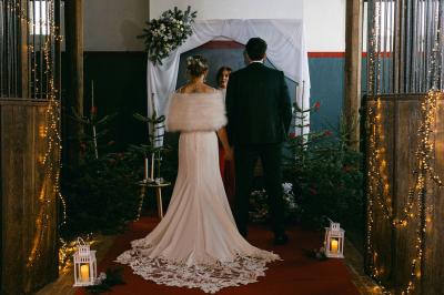 Un couple en tenue de mariage se tient devant un officiant dans un décor festif avec sapins et guirlandes lumineuses.