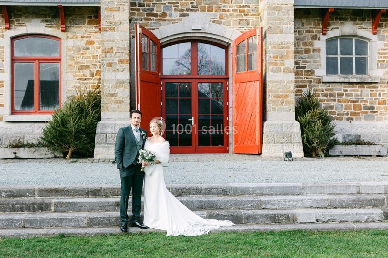 Un couple en tenue de mariage pose devant un bâtiment en pierre avec des portes rouges ouvertes.