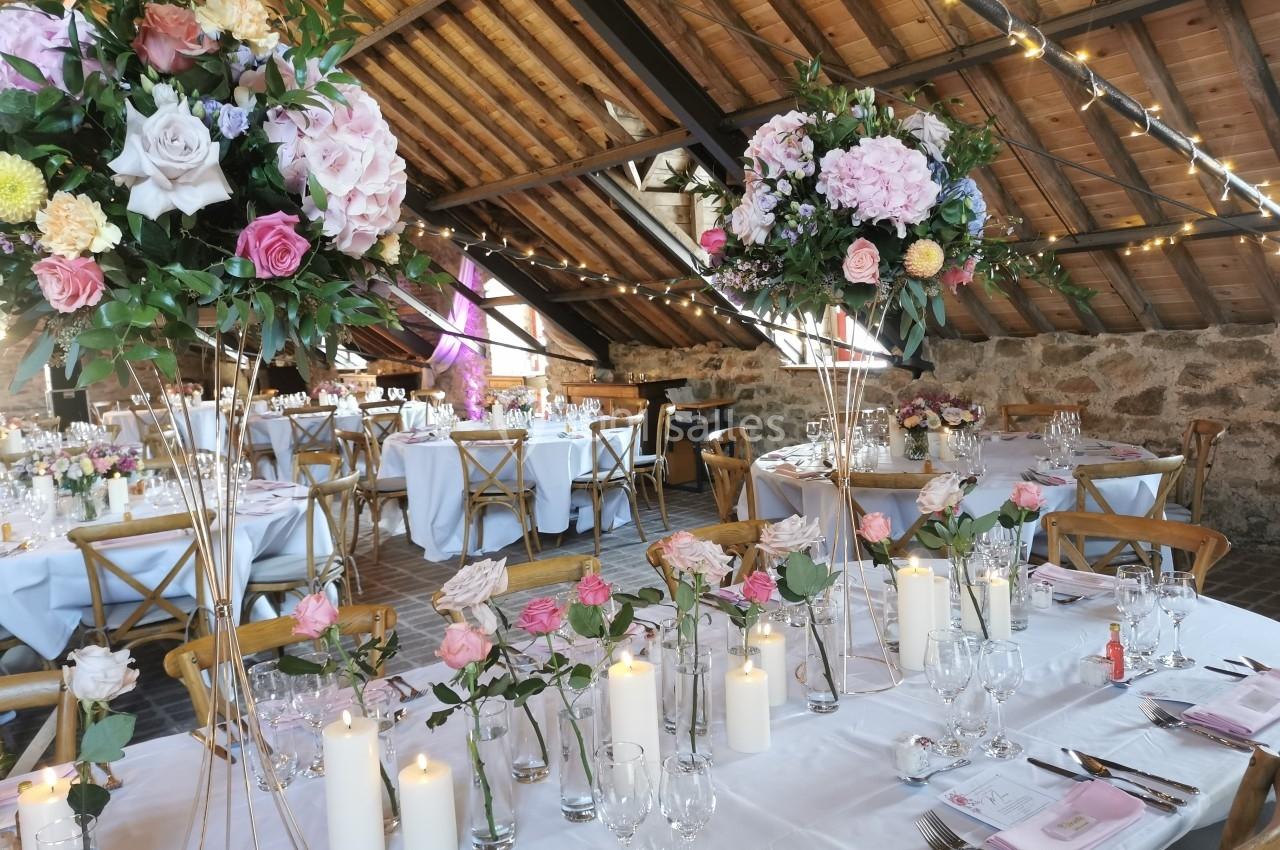 Salle de réception décorée pour un mariage, avec tables dressées, bougies et compositions florales sous un toit en bois.