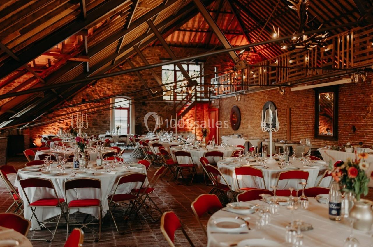 Salle de réception rustique avec tables rondes dressées, chaises rouges et poutres apparentes sous un toit en pente.