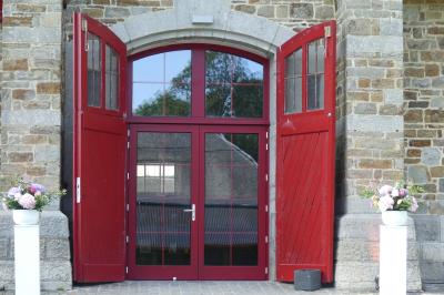 Porte d'entrée rouge à double battant ouverte, entourée d'un mur en pierre, avec des pots de fleurs de chaque côté.