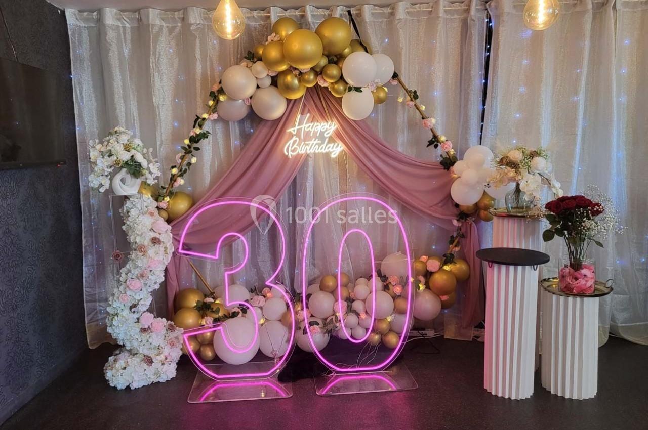 Décoration d'anniversaire avec un grand ’30’ lumineux rose, arche de ballons dorés et blancs, et fleurs sur fond scintillant.