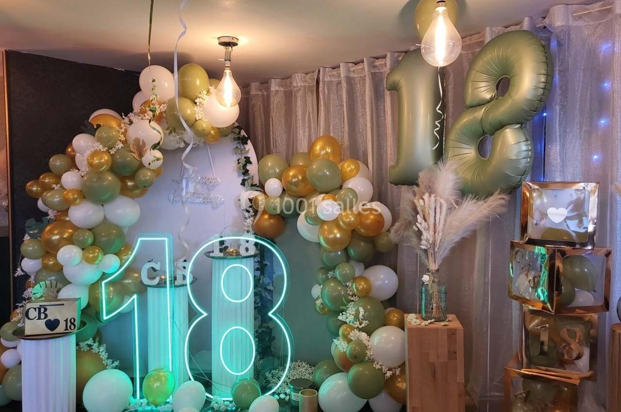 Décoration festive avec ballons dorés, blancs et verts, chiffres ’18’ lumineux et éléments décoratifs variés.