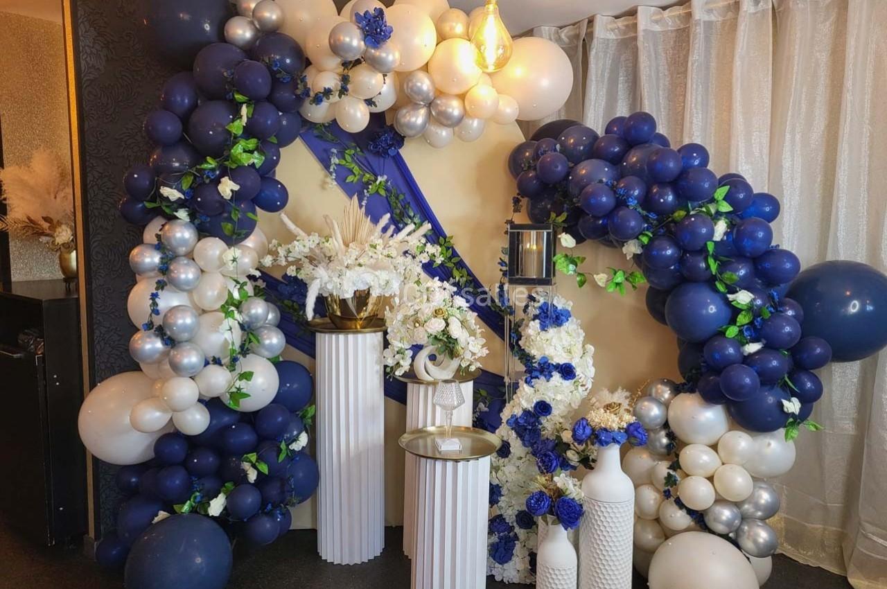 Décoration festive avec des ballons bleus, blancs et argentés entourant des colonnes ornées de fleurs blanches et bleues.