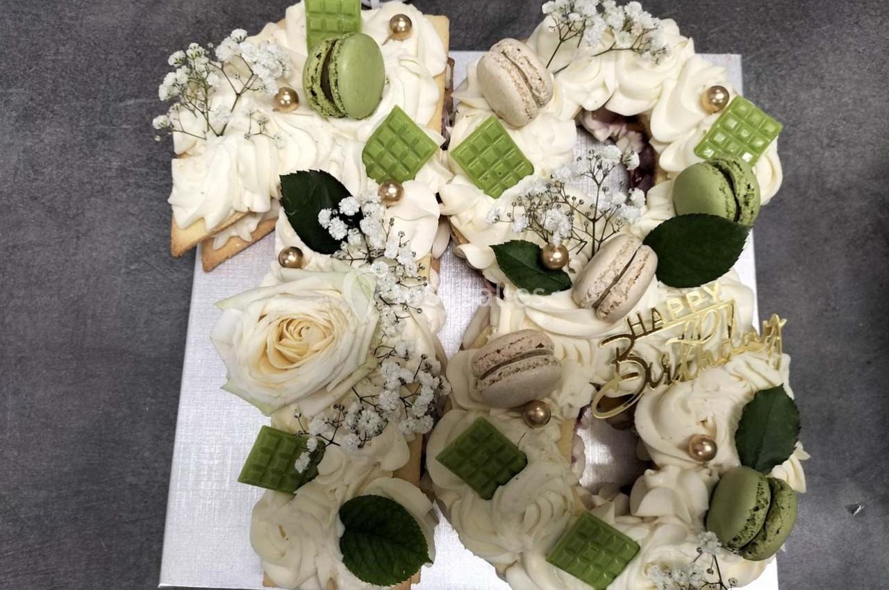 Gâteau d'anniversaire décoré de crème, macarons, chocolat vert, fleurs blanches et feuillage sur une base argentée.