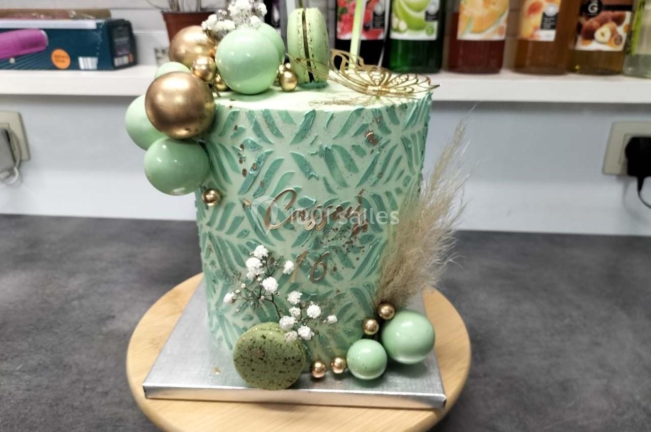 Gâteau cylindrique décoré de motifs verts, macarons, boules dorées et fleurs séchées sur un présentoir en bois.