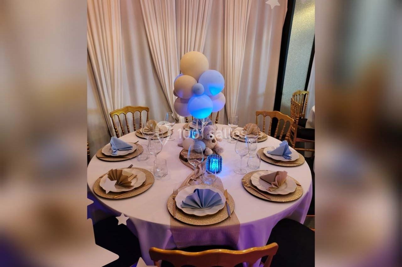 Table décorée pour un événement avec nappes blanches, serviettes pliées, vaisselle élégante et centre de table avec ballons…