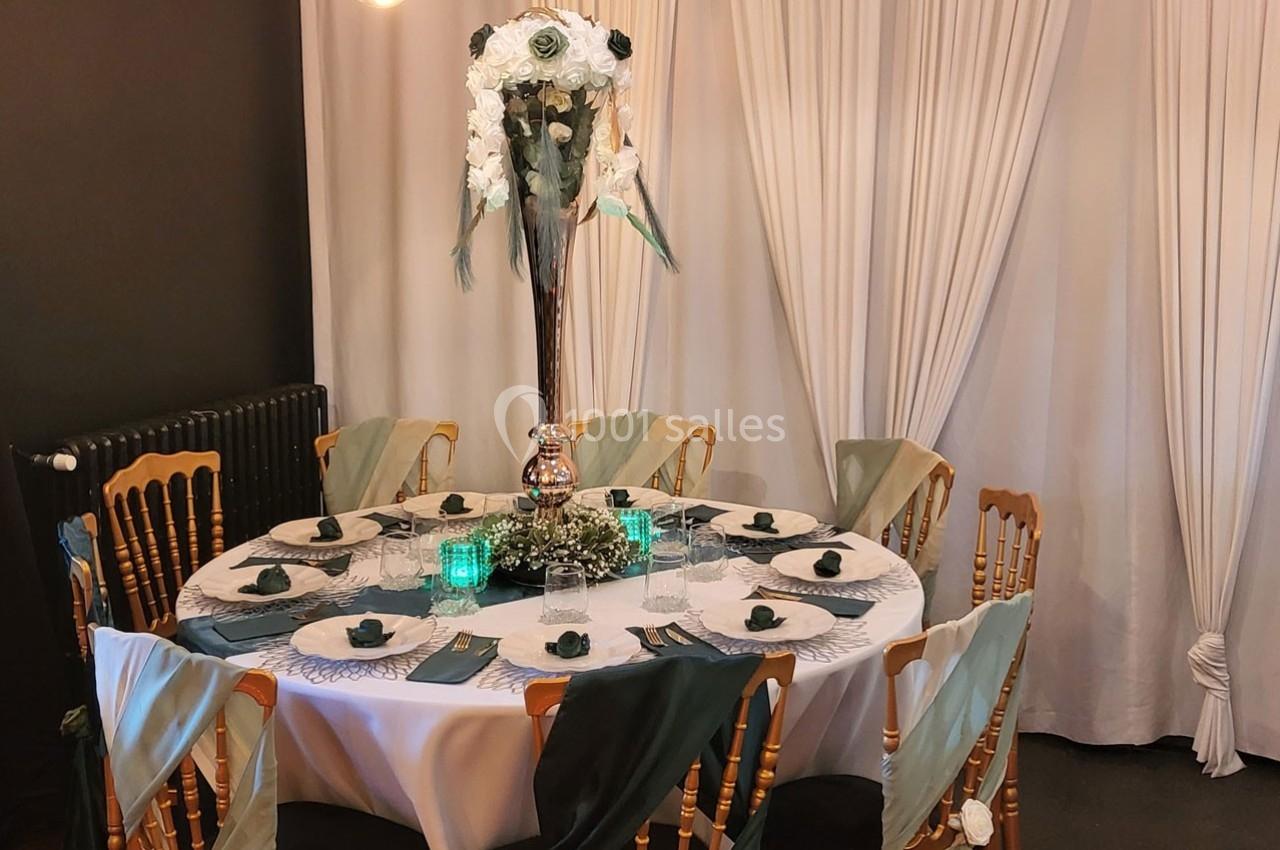 Table ronde décorée pour un événement, avec nappes blanches, chaises ornées de tissus verts et centre de table floral.