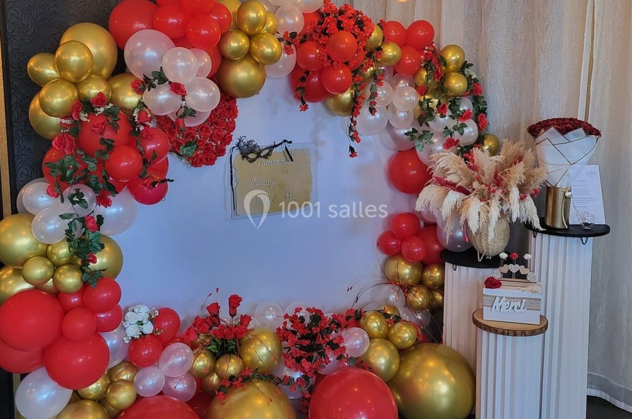 Décoration festive avec arche de ballons rouges, blancs et dorés, fleurs rouges et table avec gâteau et bouquet.