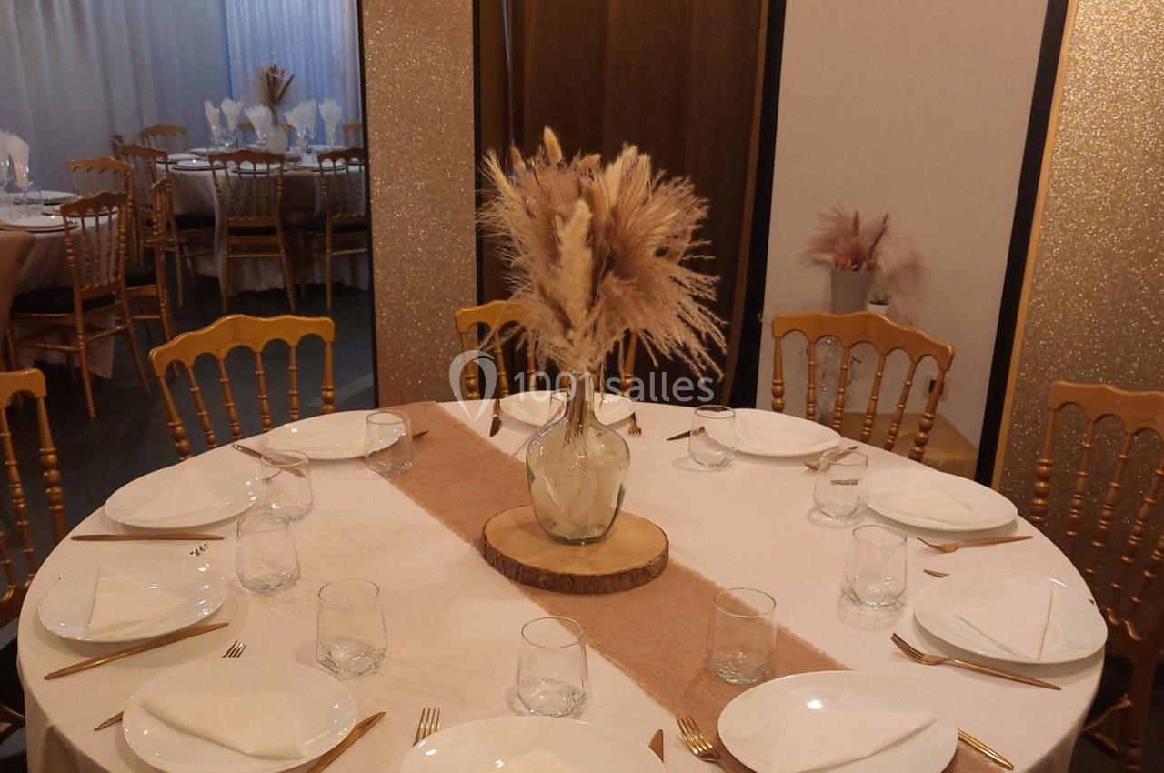 Table ronde dressée avec une nappe blanche, des assiettes, des verres, des couverts dorés et un centre de table en herbes…
