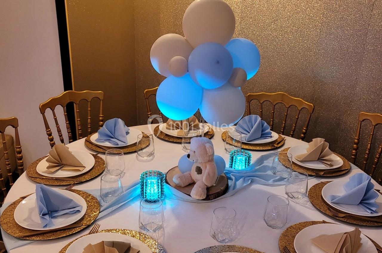 Table décorée avec des ballons bleus et blancs, un ours en peluche, serviettes pliées et assiettes dorées.
