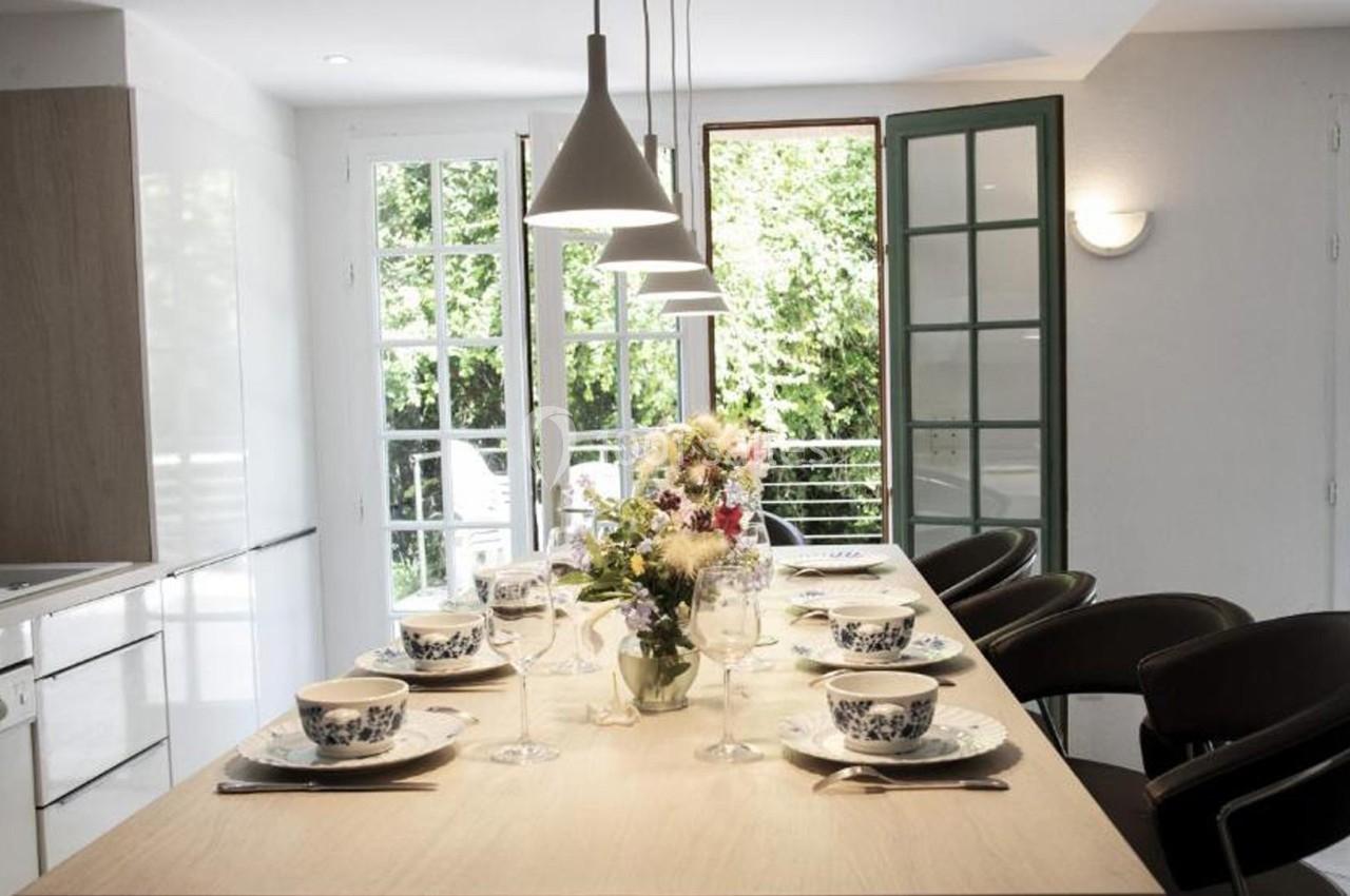 Table dressée avec vaisselle et fleurs au centre, dans une cuisine lumineuse avec grandes fenêtres donnant sur un jardin.