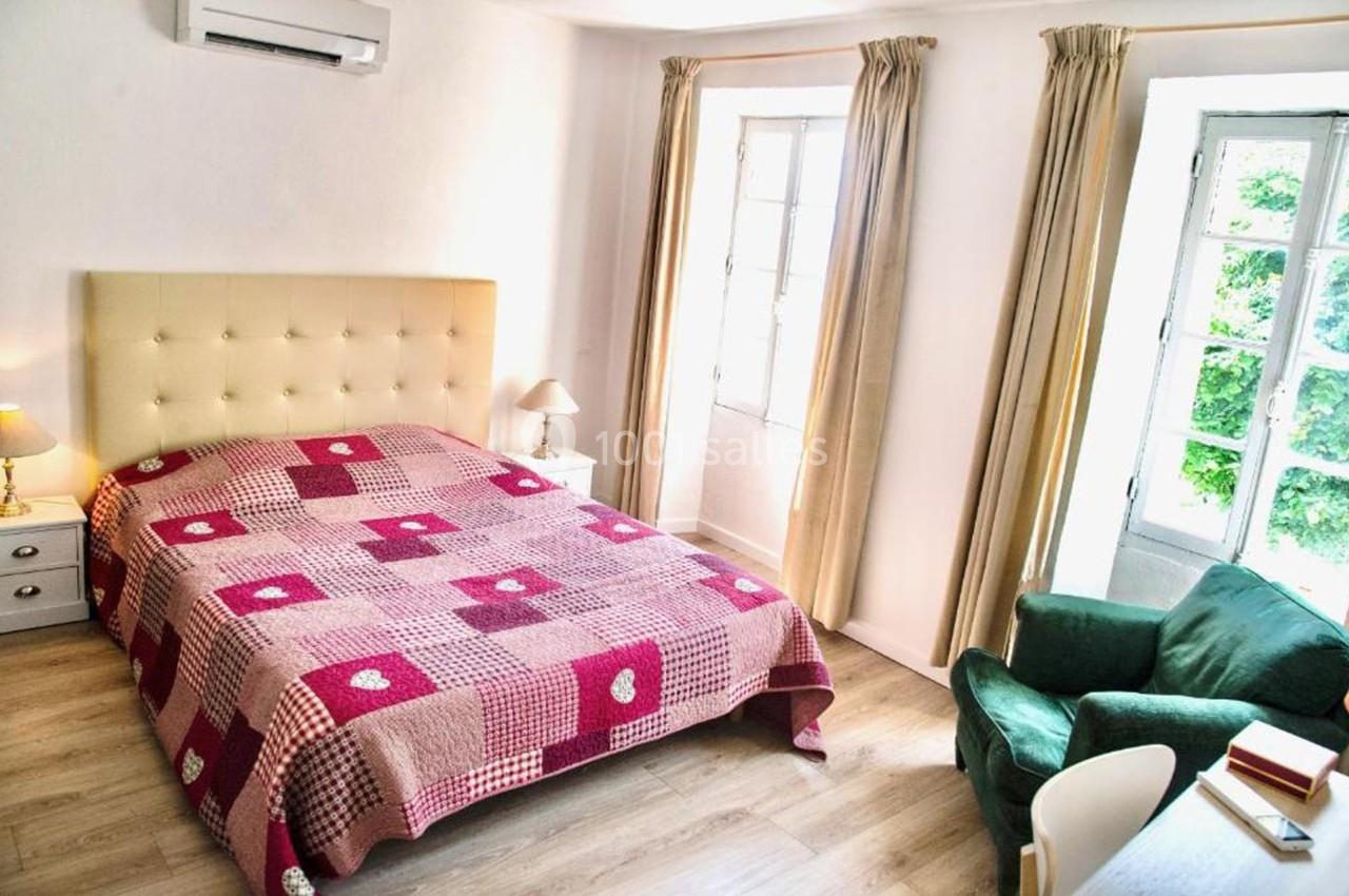 Chambre lumineuse avec lit double, couvre-lit rouge à motifs, fauteuil vert, bureau et grandes fenêtres avec rideaux beiges.