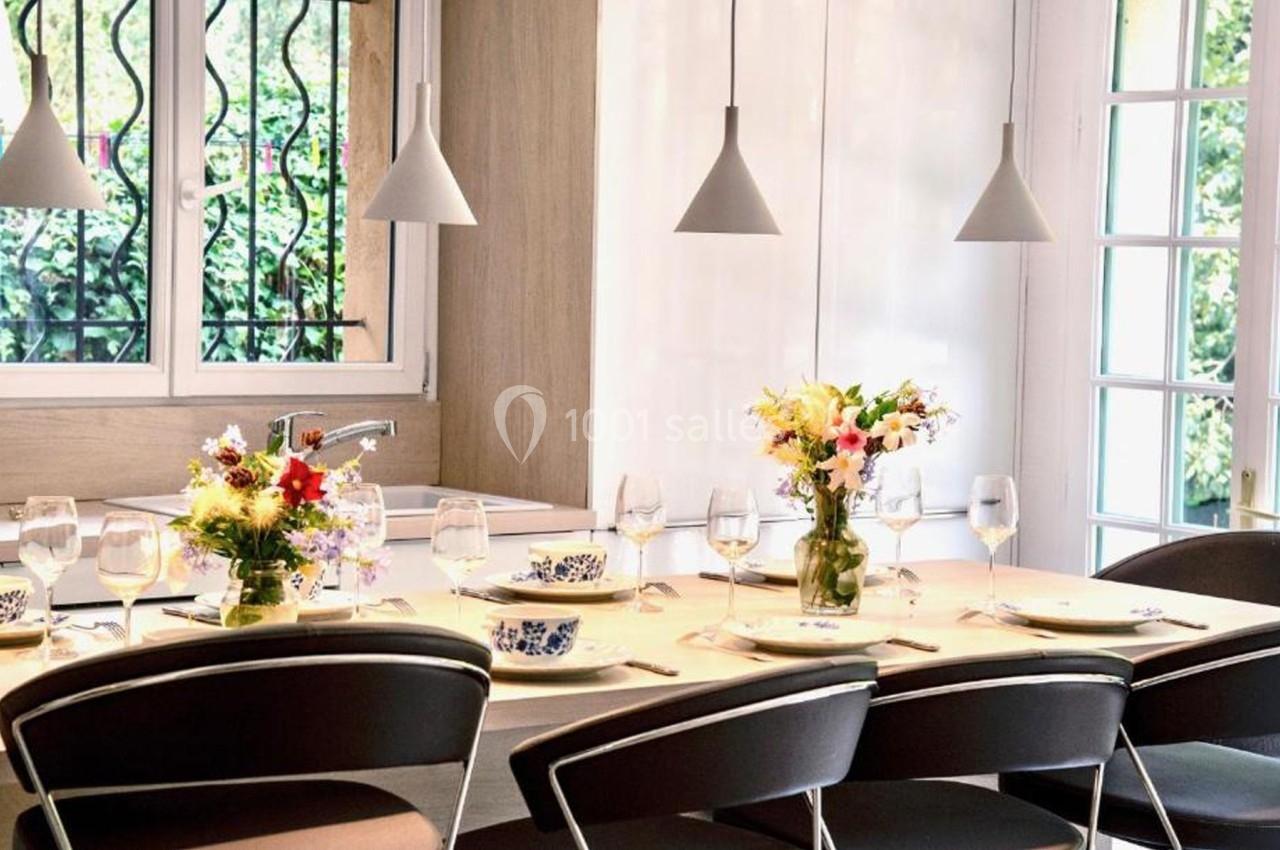 Table à manger dressée avec vaisselle et bouquets de fleurs, éclairée par des suspensions, près de grandes fenêtres.