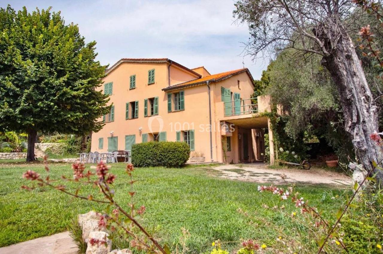 Grande maison jaune avec volets verts, entourée d'un jardin arboré et fleuri sous un ciel clair.