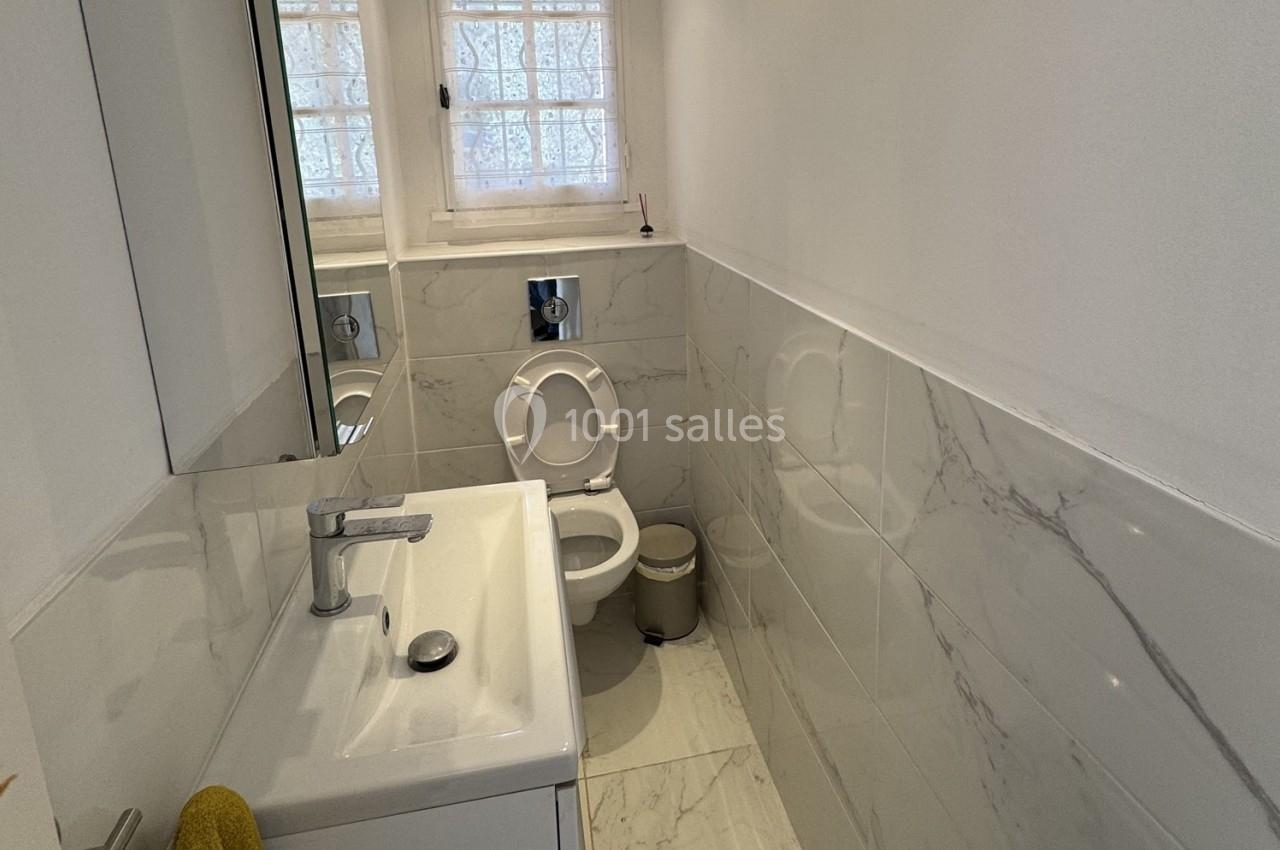 Salle de bain avec toilettes, lavabo, miroir et murs en carrelage blanc à motif marbré, éclairée par une fenêtre.