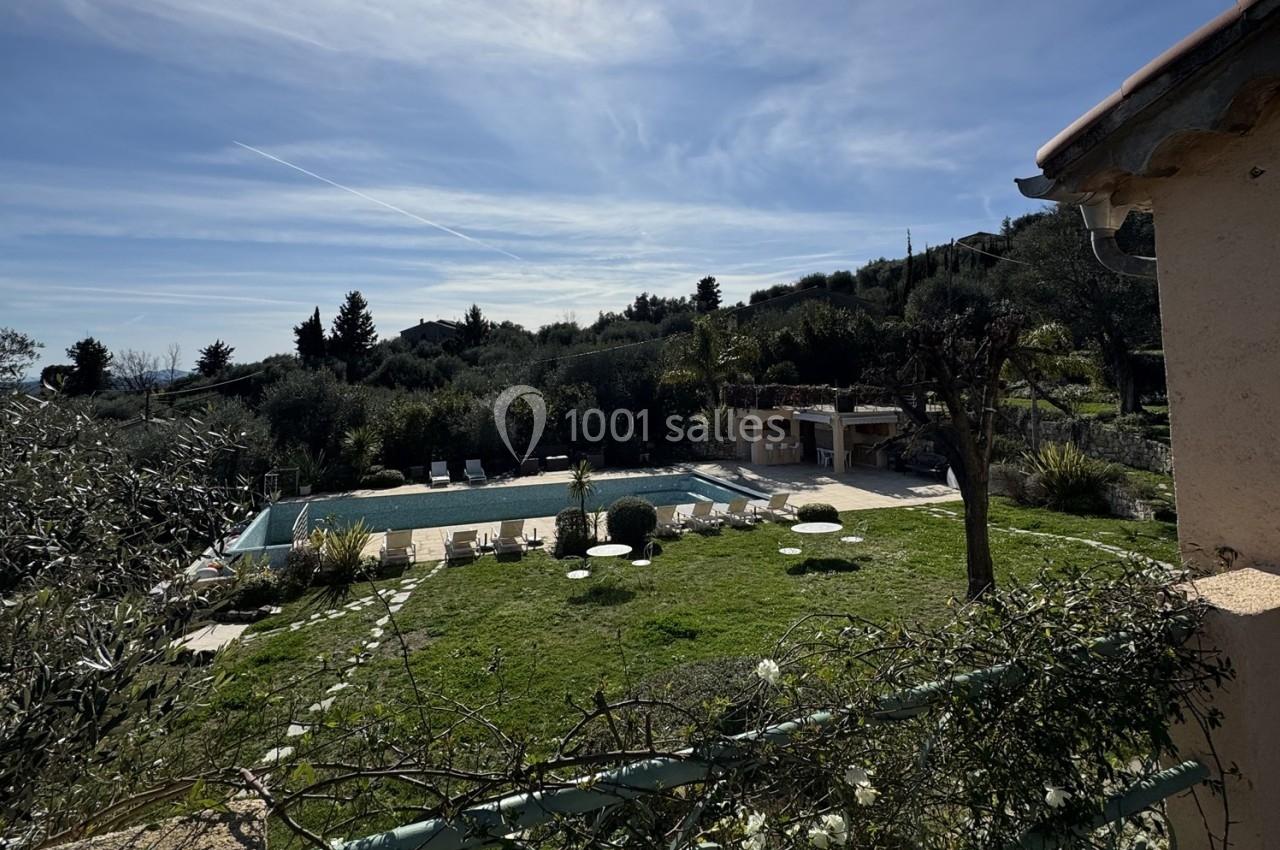 Vue d'une piscine extérieure entourée de chaises longues, pelouse et végétation, avec colline boisée en arrière-plan.
