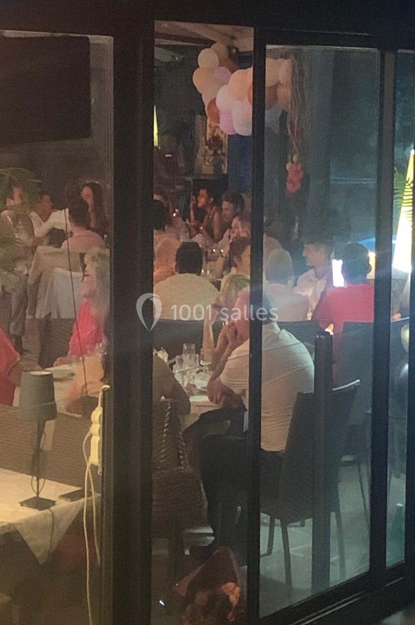 Vue à travers une vitre d'un restaurant animé en soirée, avec des convives attablés et des décorations festives.