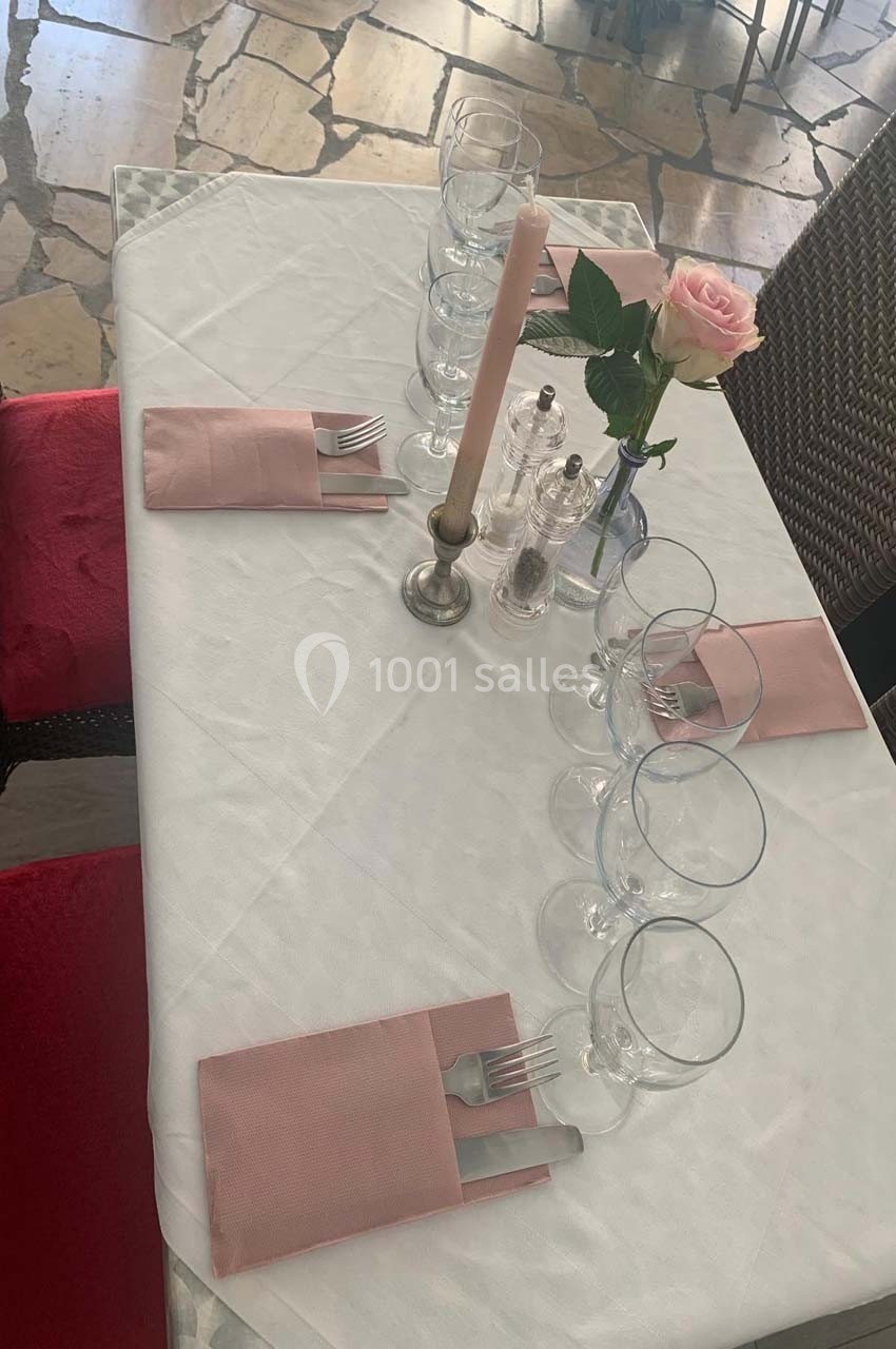 Table dressée avec nappe blanche, serviettes roses, verres, couverts, bougie et une rose dans un vase.