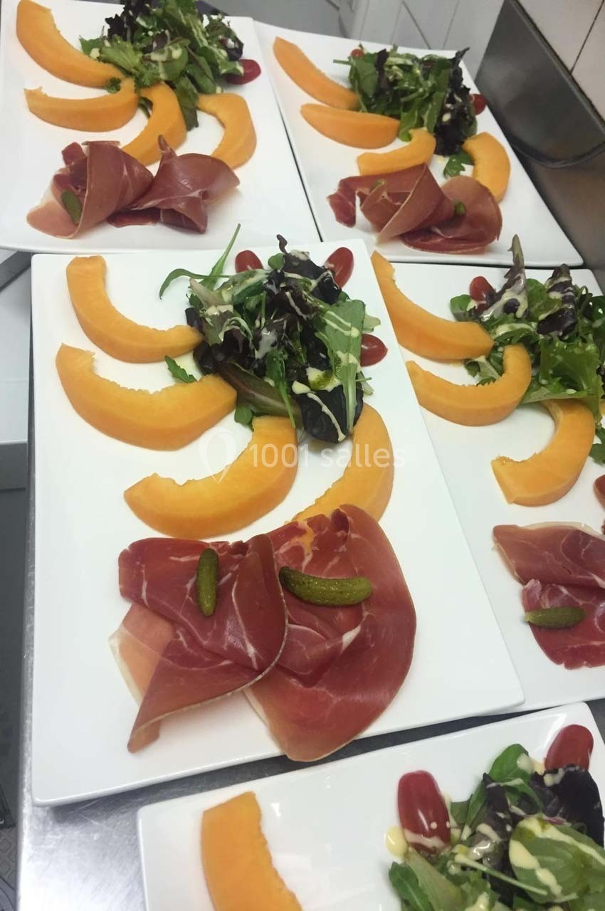 Assiettes de melon, jambon cru, cornichons et salade verte servies sur des plats blancs rectangulaires.