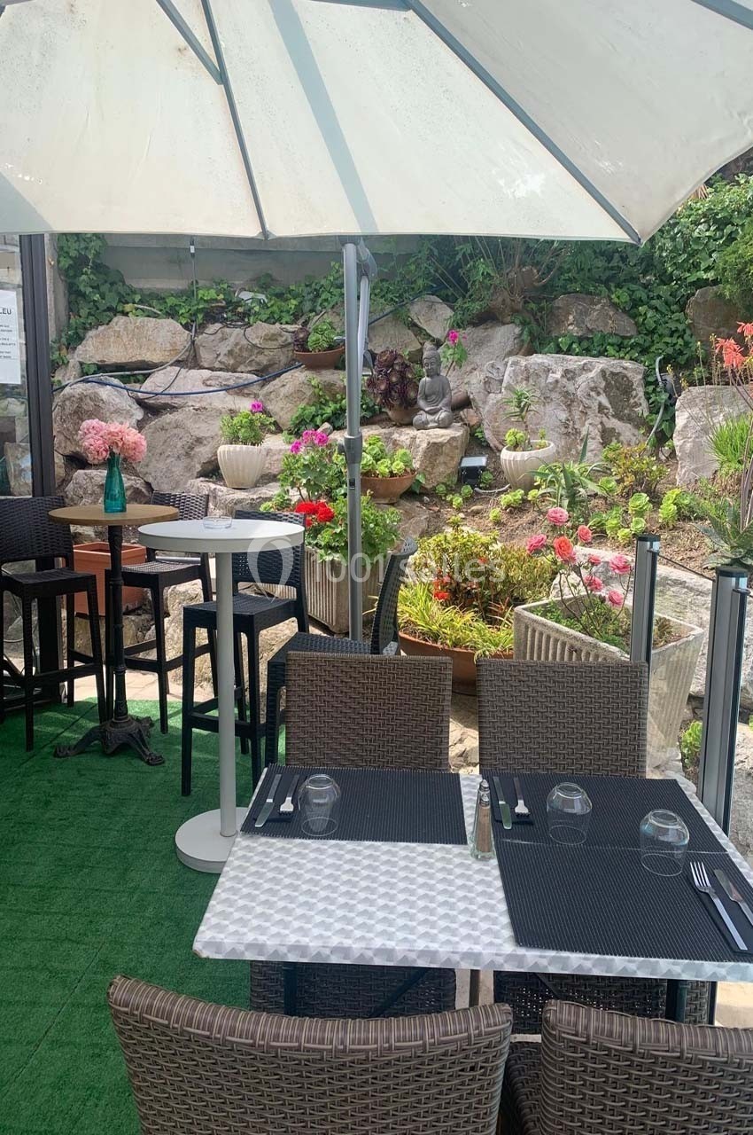 Terrasse d'un restaurant avec tables dressées, chaises en rotin, parasol blanc et jardin fleuri en arrière-plan.