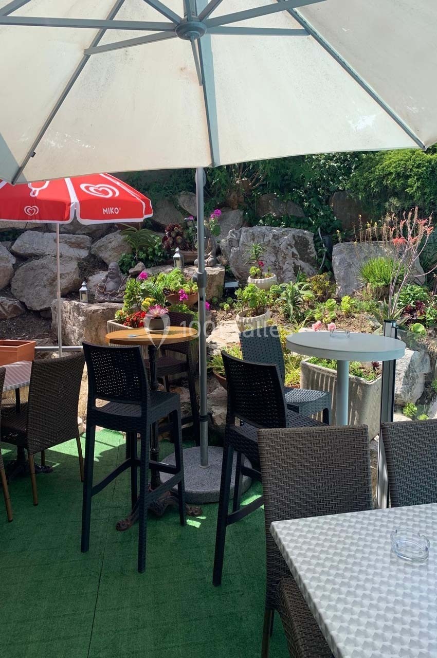 Terrasse extérieure avec tables, chaises en rotin, parasols et décor floral entouré de rochers.